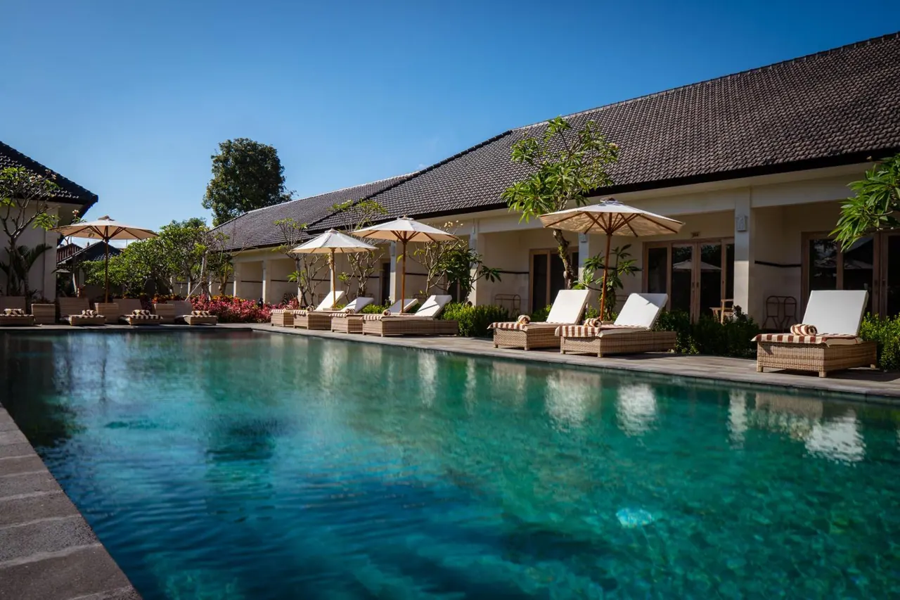 A long swimming pool with white-cushioned wicker sun loungers and parasols along its edge, beside a single-storey building with a tiled roof.