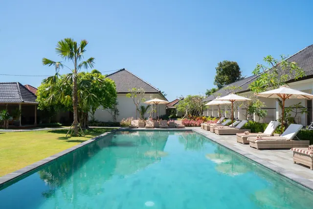 A long rectangular swimming pool with turquoise water runs past sun loungers and white parasol umbrellas beside a lawn and low white buildings under a clear blue sky.