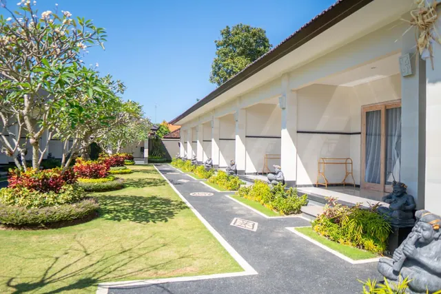 A paved path runs between a lawn with frangipani trees and shrubs, beside a covered walkway of guest room doors with stone statues along the edge.