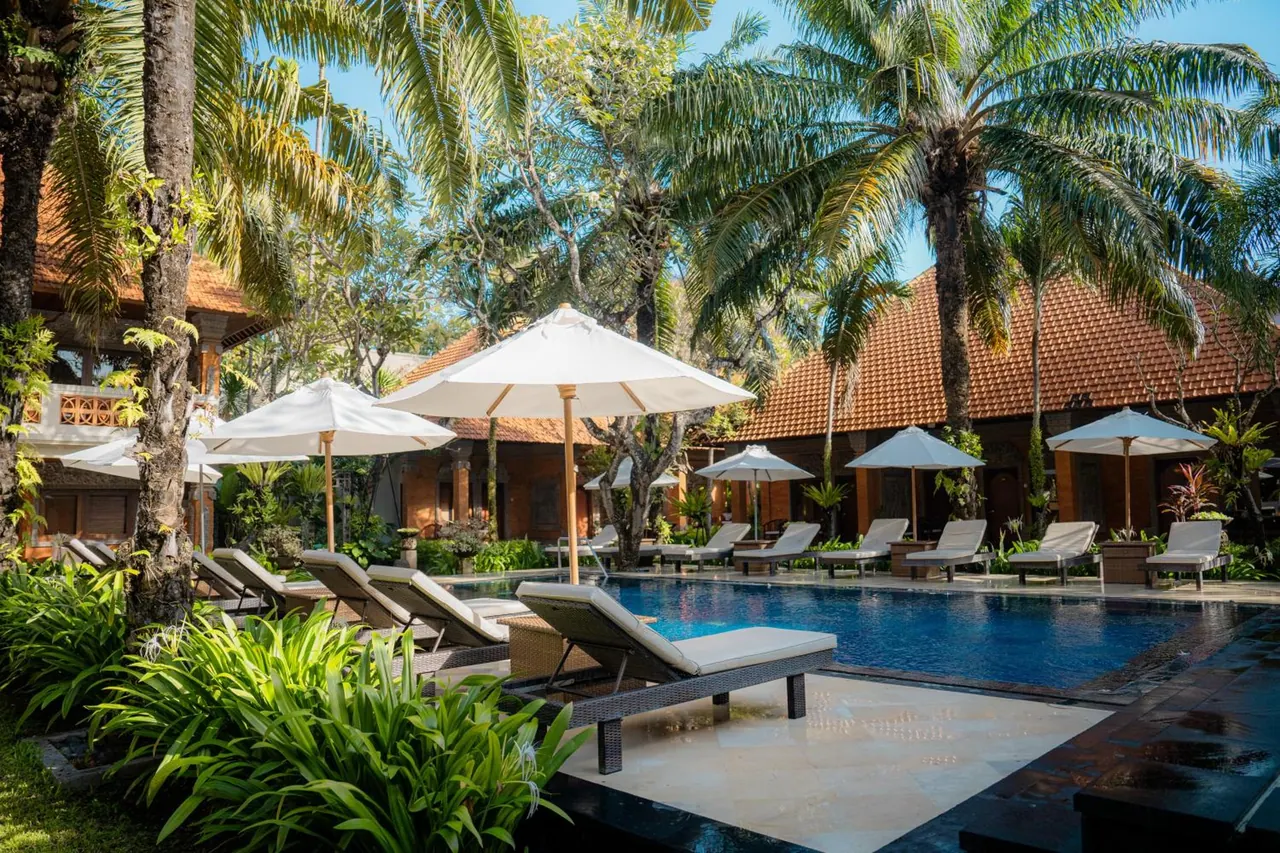 A swimming pool with white sun loungers and white parasol umbrellas beside it, surrounded by palm trees and terracotta-roofed buildings.