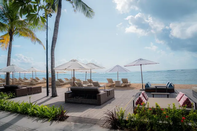 A wooden deck with wicker daybeds, sun loungers and parasols faces a sandy beach and the sea under a partly cloudy sky.
