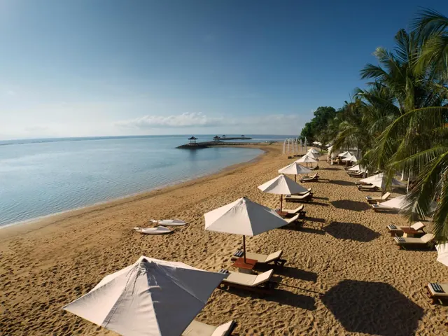A sandy beach with rows of sun loungers and white umbrellas runs along calm blue water, with palm trees on the right and a small pavilion on a causeway in the distance.