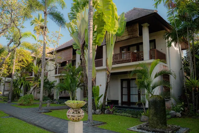 A two-storey Balinese-style villa with a tiled roof and wooden balcony railings stands among palm trees and tropical plants, with a stone pathway and a planter urn in the foreground.