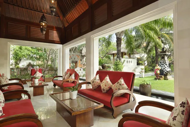 A traditional open lounge with red sofas, floral cushions and a glass coffee table, with large windows looking onto a palm-lined garden and a stone statue.