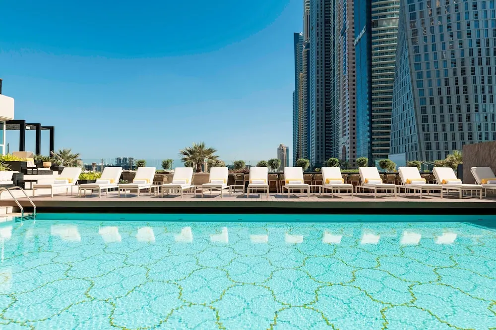 A rooftop swimming pool with white sun loungers along the deck, with tall towers and a city skyline behind.