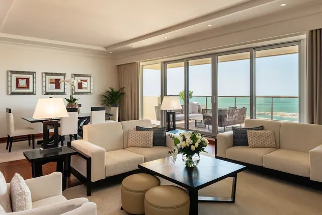 A living room with two cream sofas, a coffee table with white flowers, and a dining area, with floor-to-ceiling glass doors opening onto a balcony with a view of the sea.