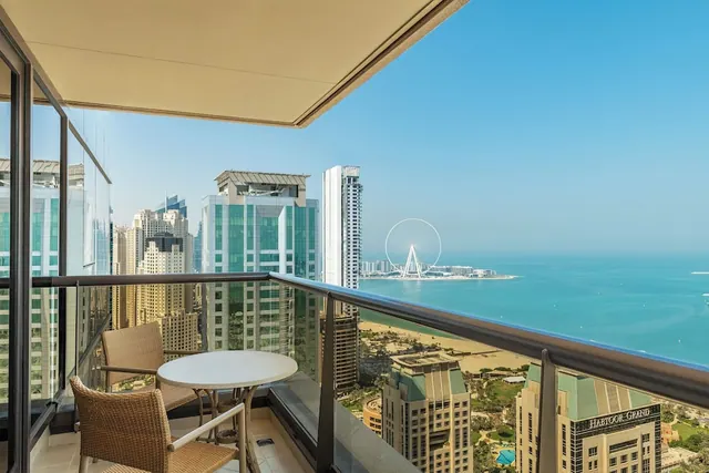 A furnished balcony with two wicker chairs and a round table overlooks the sea, a sandy beach, high-rise buildings and the Ain Dubai ferris wheel, with the Habtoor Grand hotel building below.