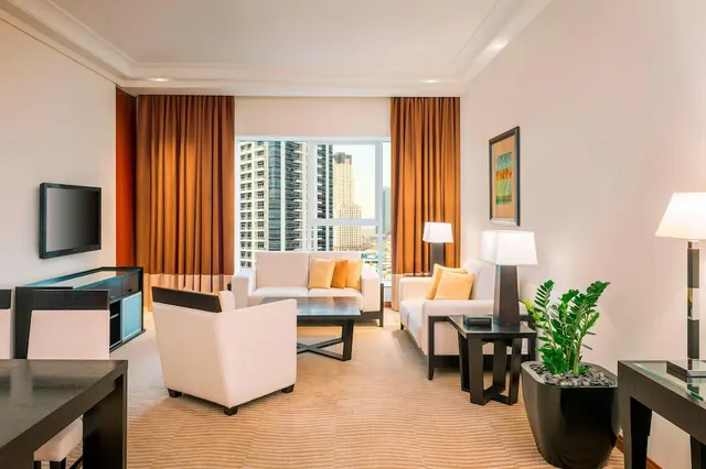 A furnished living area with white armchairs, a sofa with yellow cushions, a wall-mounted television, and high-rise towers visible through the window.