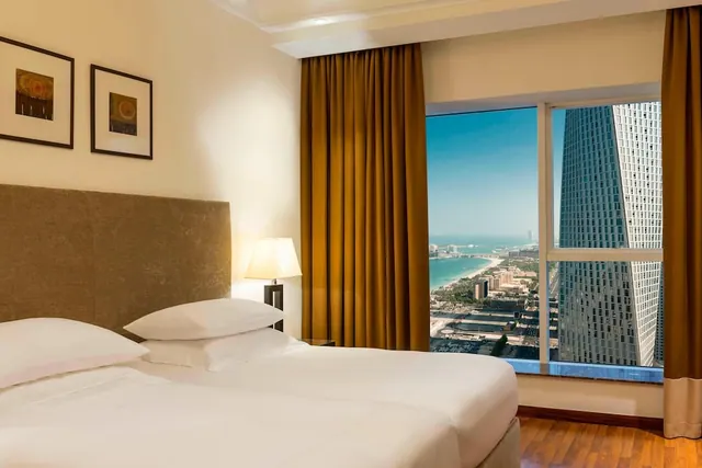 A bedroom with a made bed, framed artwork, gold curtains and a large window overlooking the sea, a sandy beach and a tall glass building.