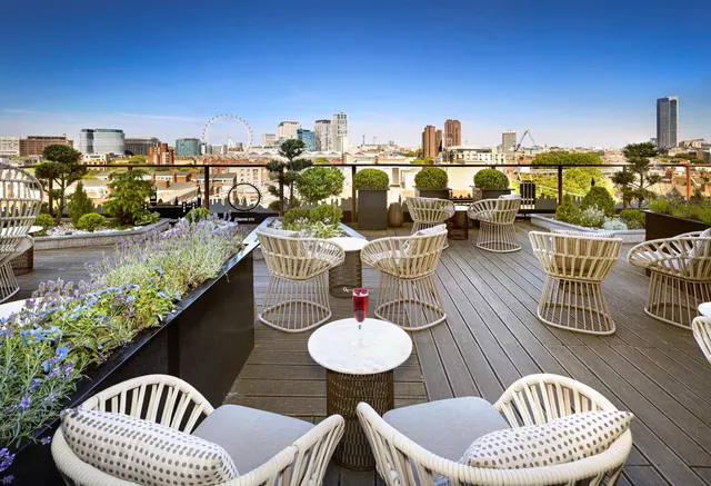 A rooftop terrace with wicker armchairs, a round marble table holding a drink, lavender planters and a view across London that includes the London Eye.