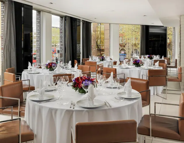 A dining room with round tables set with white linen, glassware and napkins, with tall windows looking onto a street with brick buildings and trees.