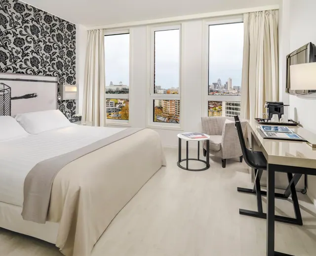 A hotel bedroom with a king bed, a black and white patterned feature wall, a desk with a coffee machine and a wall-mounted TV, and windows showing a city skyline.