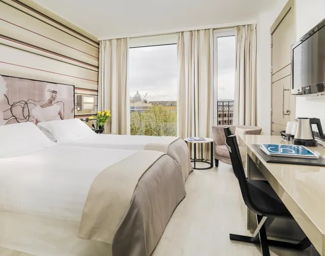 A hotel bedroom with two beds, a desk and chair, a wall-mounted television, and a window looking out over a city with a domed building in the distance.