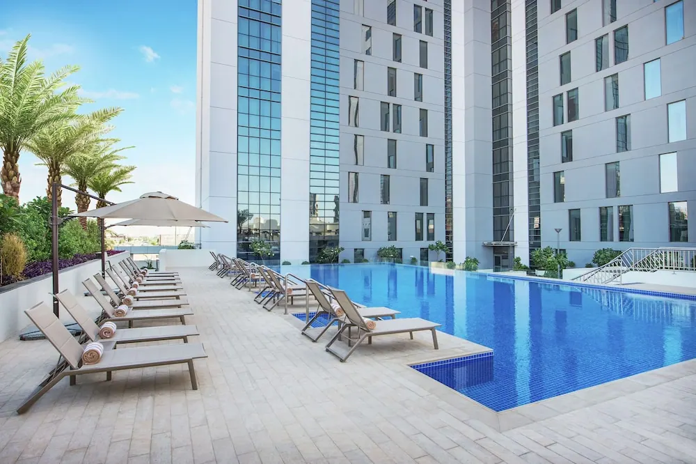 An outdoor pool with blue tiled edges sits beside a sunny deck lined with sun loungers, a shade umbrella, and tall white hotel towers behind it.