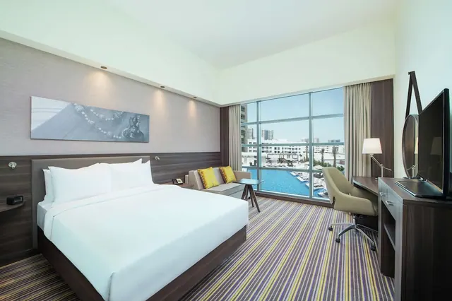 A hotel bedroom with a made king bed, a seating area with yellow cushions, a desk and television, and a large window overlooking a pool and buildings.