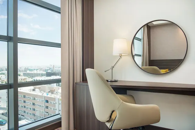 A work desk with a cream chair, a table lamp and a round mirror sits beside floor-to-ceiling windows overlooking low-rise city buildings.