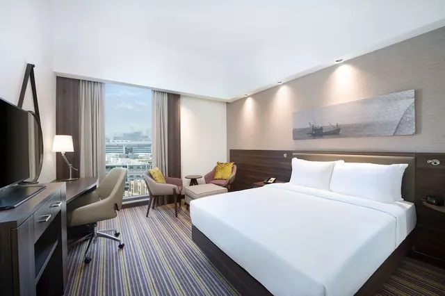 A bedroom with a large white bed, two armchairs by the window, a desk and a wall-mounted television, with city buildings visible through the window.