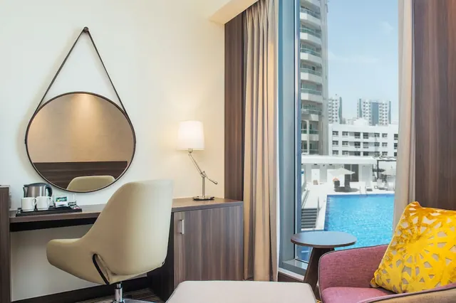 A hotel room with a desk, round mirror, lamp and kettle, and a window overlooking a swimming pool and residential towers.