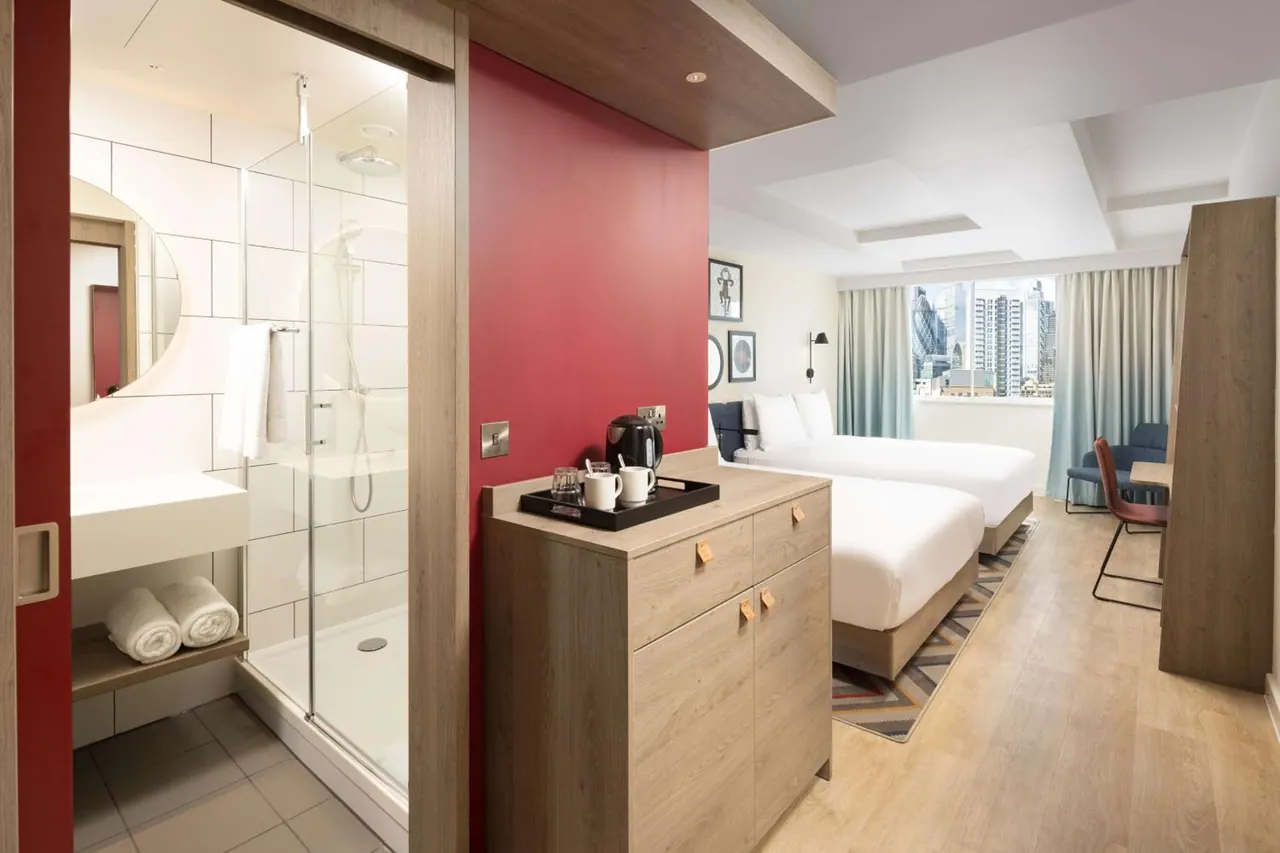 A hotel room with two beds, a tea and coffee tray on a wooden cabinet, and a walk-in shower visible through a glass door, with a window view of the Gherkin and city towers.