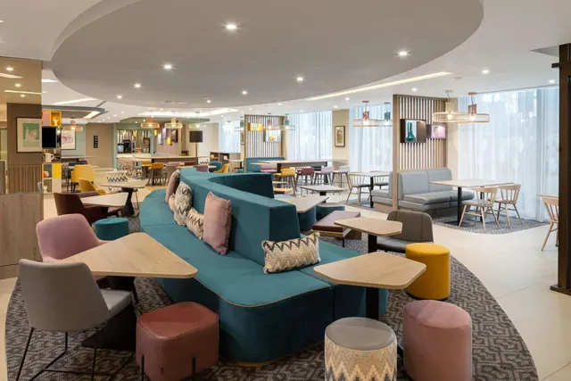 A lounge area with a curved teal sofa, low wooden tables, patterned stools and grey seating, with floor-to-ceiling windows covered by sheer curtains.