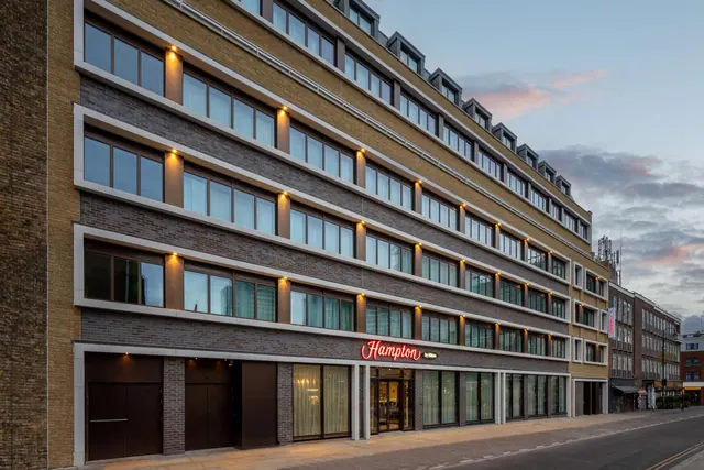 The exterior of a Hampton by Hilton hotel, a multi-storey brick building with a red illuminated sign above the entrance, on a city street at dusk.