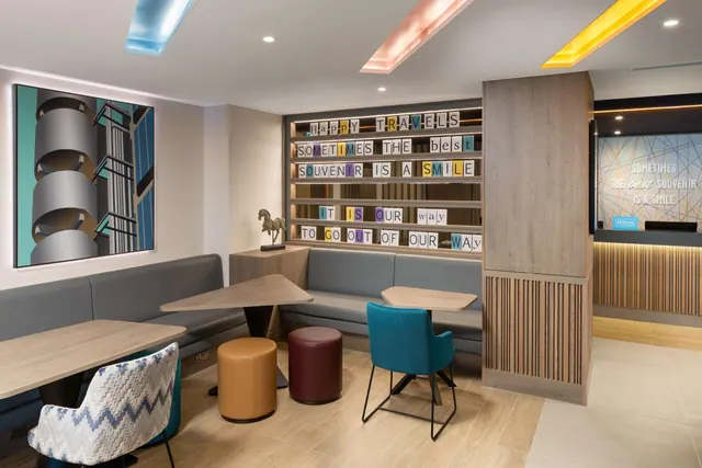 A lounge seating area with grey booth benches, wooden tables and coloured stools, beside a wall of letter tiles reading happy travels and a reception counter with a Hilton logo.