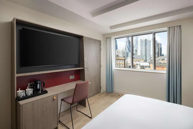 A hotel bedroom with a wall-mounted television, a desk with a kettle and mugs, and a window showing city buildings including the Gherkin.