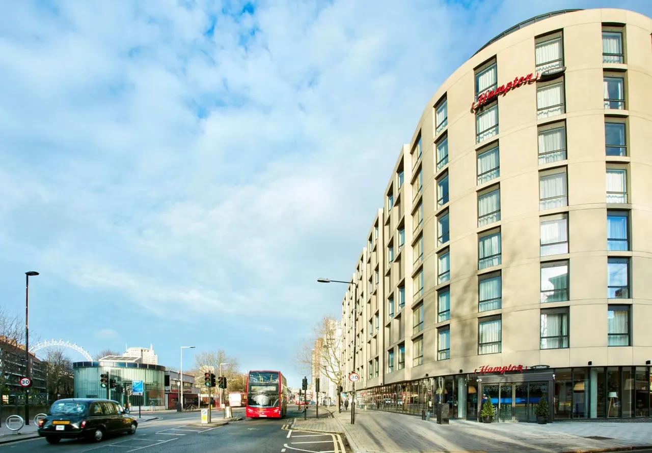 A curved hotel facade with the Hampton name on it stands beside a street with a black taxi and a red double-decker bus, with the London Eye partly visible at left.