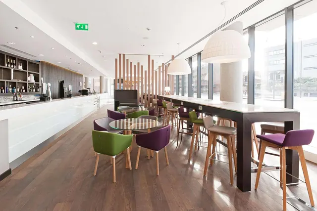 A long bar counter with bottle shelves sits beside a lounge area with green and purple armchairs, round glass tables, high stools and pendant lamps, with large windows onto a street.