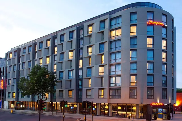 The exterior of a multi-storey hotel building at dusk, with lit windows, red Hampton neon signs, a street tree and traffic lights in front.