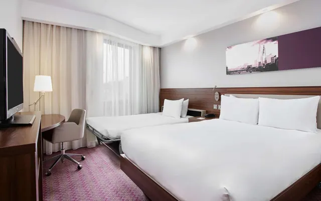 A hotel bedroom with a large double bed, a pull-out extra bed, a desk with a television, and a framed print of a London skyline on the wall.