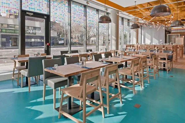 A restaurant dining room with wooden tables and chairs, a teal floor, pendant lamps, and large windows looking onto a street.