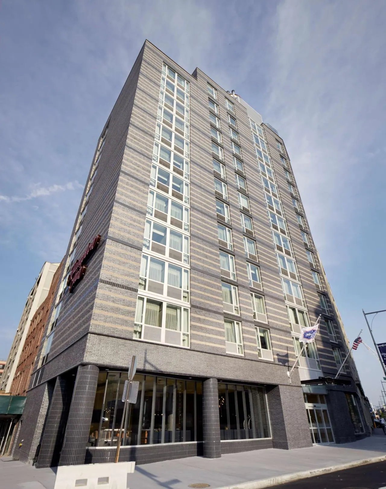 Low-angle view of a tall brick hotel building with a red logo sign at street level under a partly cloudy blue sky.