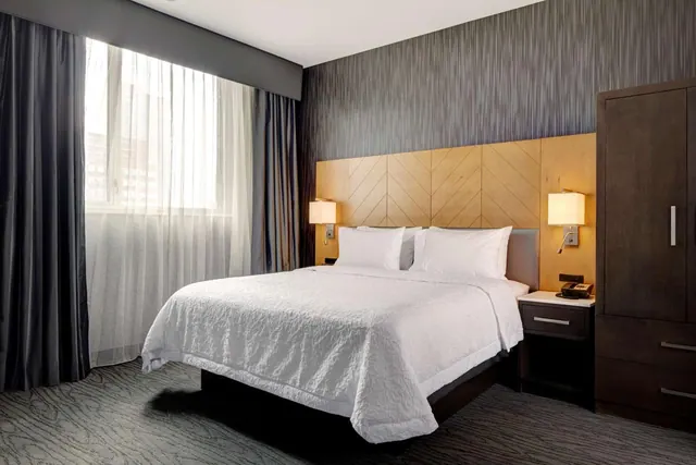 A bedroom with a made king bed with white bedding, a wood-panelled headboard, bedside lamps, a dark wooden wardrobe, and a window with sheer curtains.