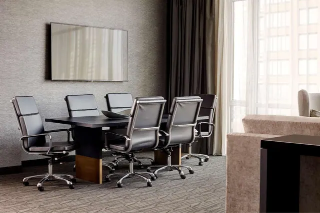 A dark table with six black leather office chairs stands in a room with a wall-mounted television, a sofa on the right, and a window with sheer curtains.