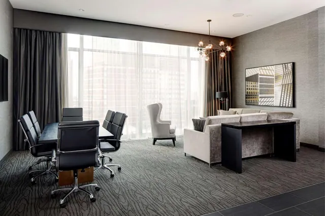 A suite living area with a dark table and black leather chairs, a grey sofa, a wingback armchair, and floor-to-ceiling windows looking onto city buildings.