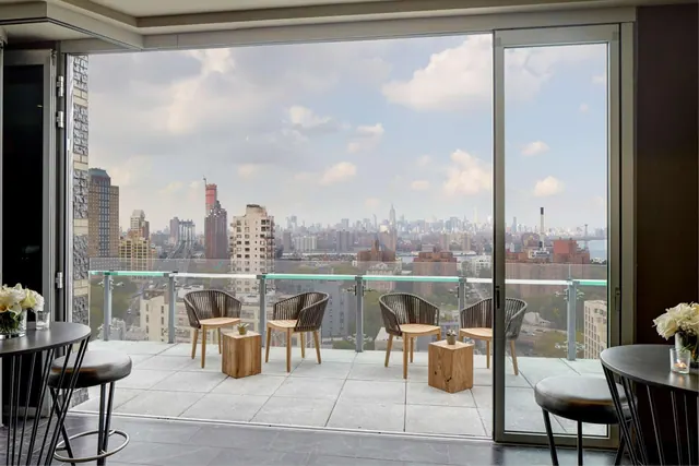 A terrace with wooden chairs and small tables sits beyond floor-to-ceiling glass doors, with a view of the Manhattan skyline including the Empire State Building.