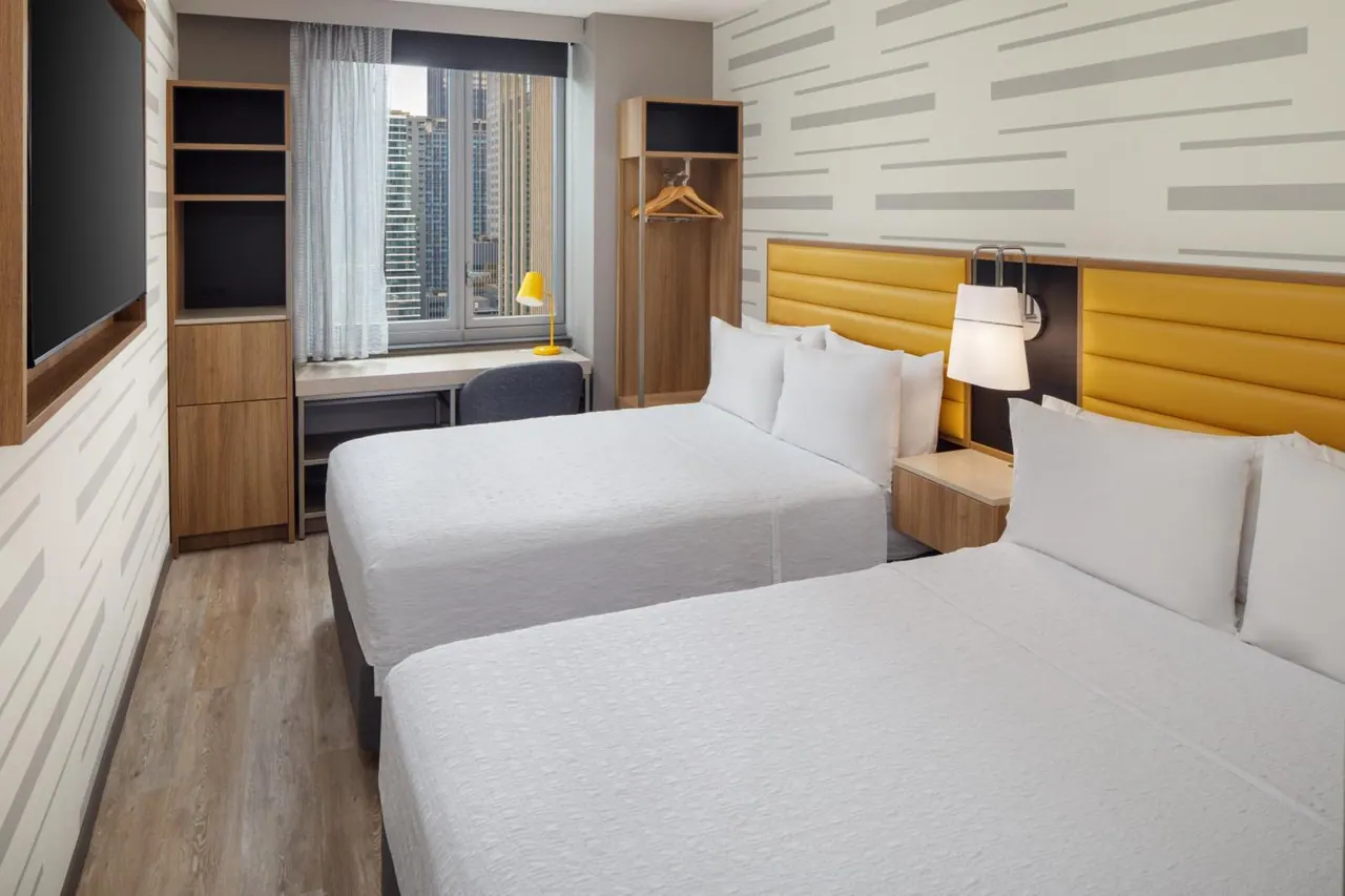 A hotel room with two beds with yellow headboards, a desk, a wardrobe, a wall-mounted television, and a window showing tall city buildings.