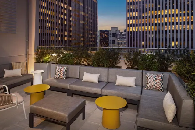 A rooftop terrace with grey sectional sofas, yellow side tables and planters, with lit office towers and city buildings visible behind at dusk.