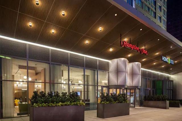 Night exterior of a hotel entrance canopy with a red Hampton Inn neon sign, a Home2 Suites sign, ceiling downlights, glass lobby frontage and planters along the pavement.