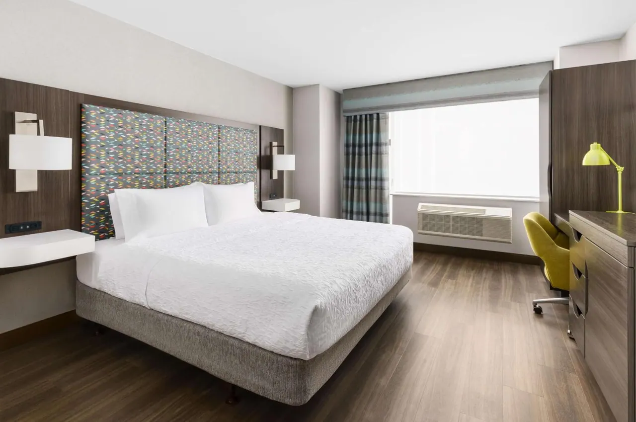 A king bed with white linens sits across from a desk with a yellow chair, a wall-mounted air conditioning unit under a window, and a wooden dresser by the wall.