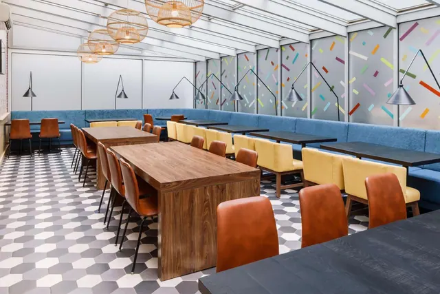 A restaurant dining room with blue banquette seating, wooden tables, orange leather chairs and a hexagonal tiled floor under a glass roof.