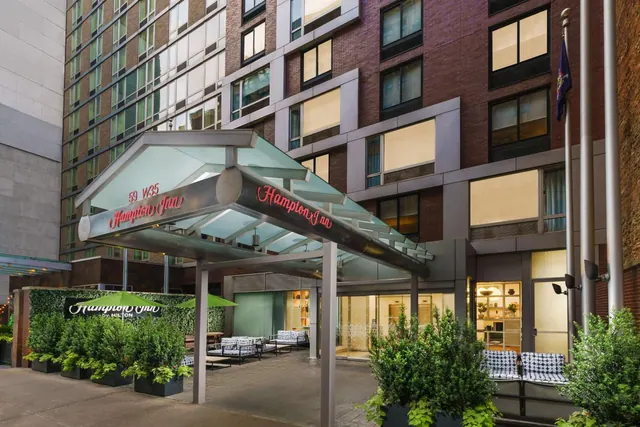 Exterior of a Hampton Inn entrance with a glass canopy, potted shrubs, outdoor benches and a brick and glass facade at dusk.