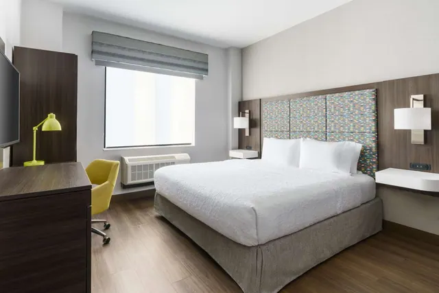 A bedroom with a king bed, a colourful upholstered headboard, a window with a roller blind, a wall-mounted air conditioner unit, a wooden desk with a yellow chair and a television.