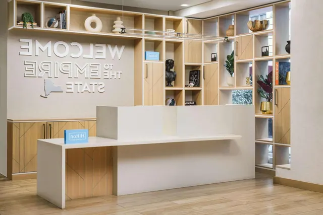 The lobby reception desk stands in front of a wall reading Welcome to the Empire State, with shelves of decor and Hilton Honors signs on the counter.