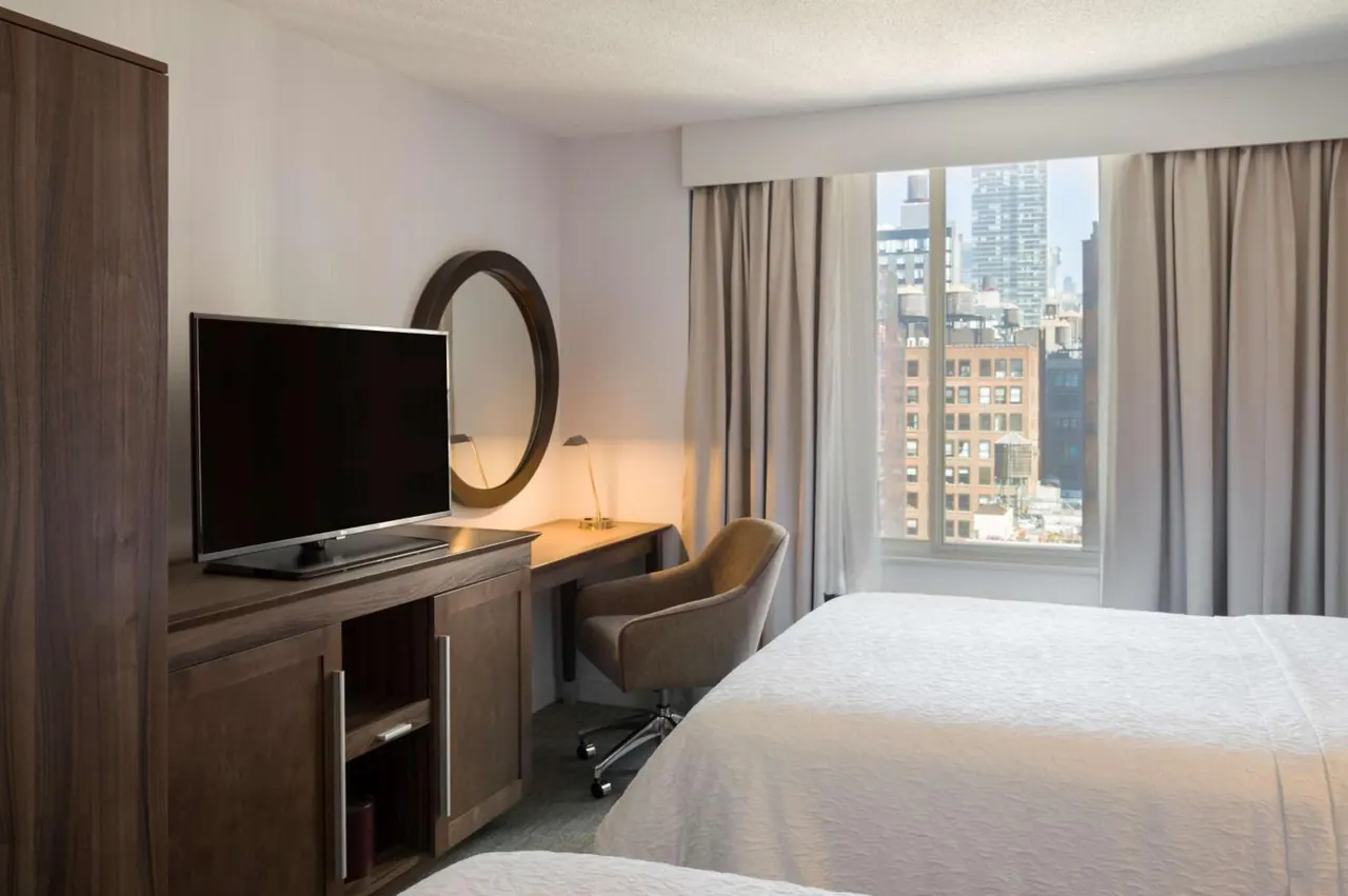 A hotel bedroom with a double bed, a flat-screen television on a wooden cabinet, a round mirror, a desk with a chair, and a window looking out over city buildings.