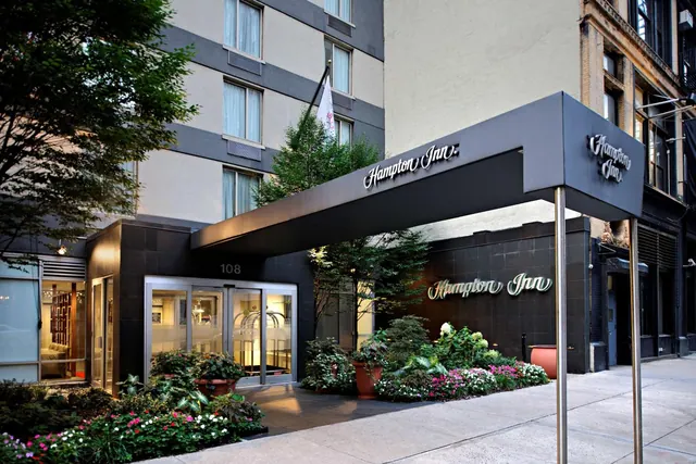 The entrance of a Hampton Inn hotel with a black canopy, glass doors, and flowering plants along the sidewalk, photographed at dusk.