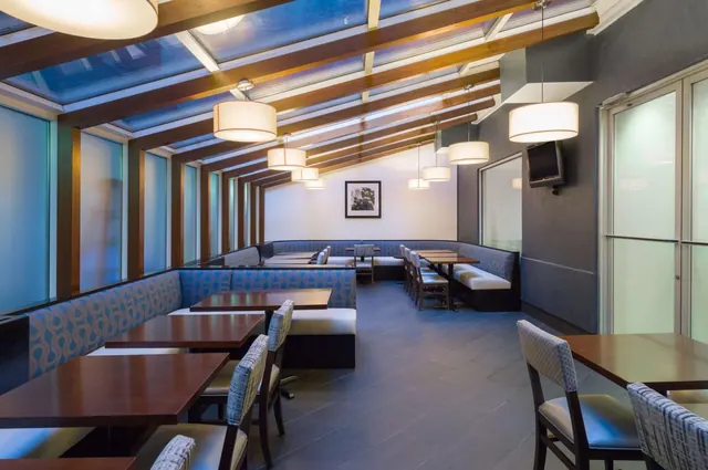 An empty restaurant dining room with blue upholstered banquettes, wooden tables, pendant lamps and a skylit ceiling with timber beams.