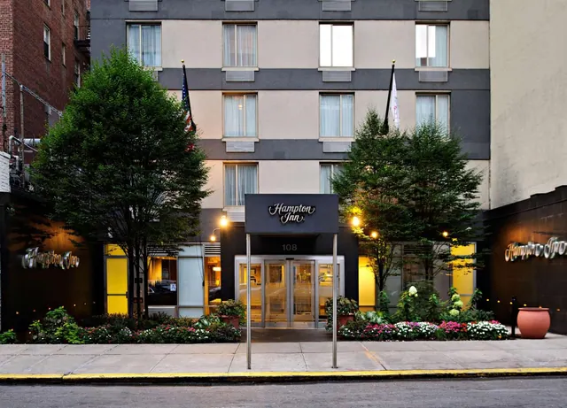 Front exterior of a Hampton Inn building at dusk, showing the entrance awning, illuminated windows, trees and flowerbeds along the sidewalk.