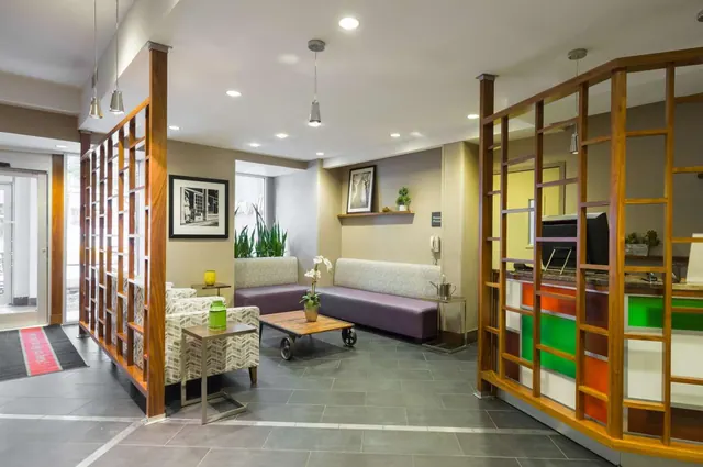 A hotel lobby lounge with purple banquette seating, patterned armchairs and a wooden coffee table, with a reception desk behind wooden screens on the right.